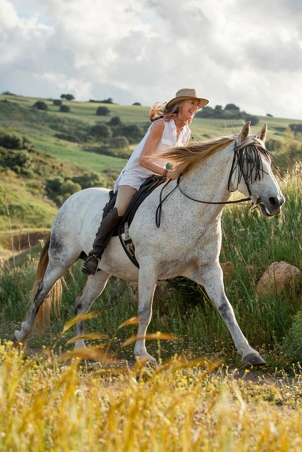 A woman riding a horse on a scenic trail through the countryside or a ranch, surrounded by lush green fields, trees, and blue sky, with the horse's mane flowing in the wind.